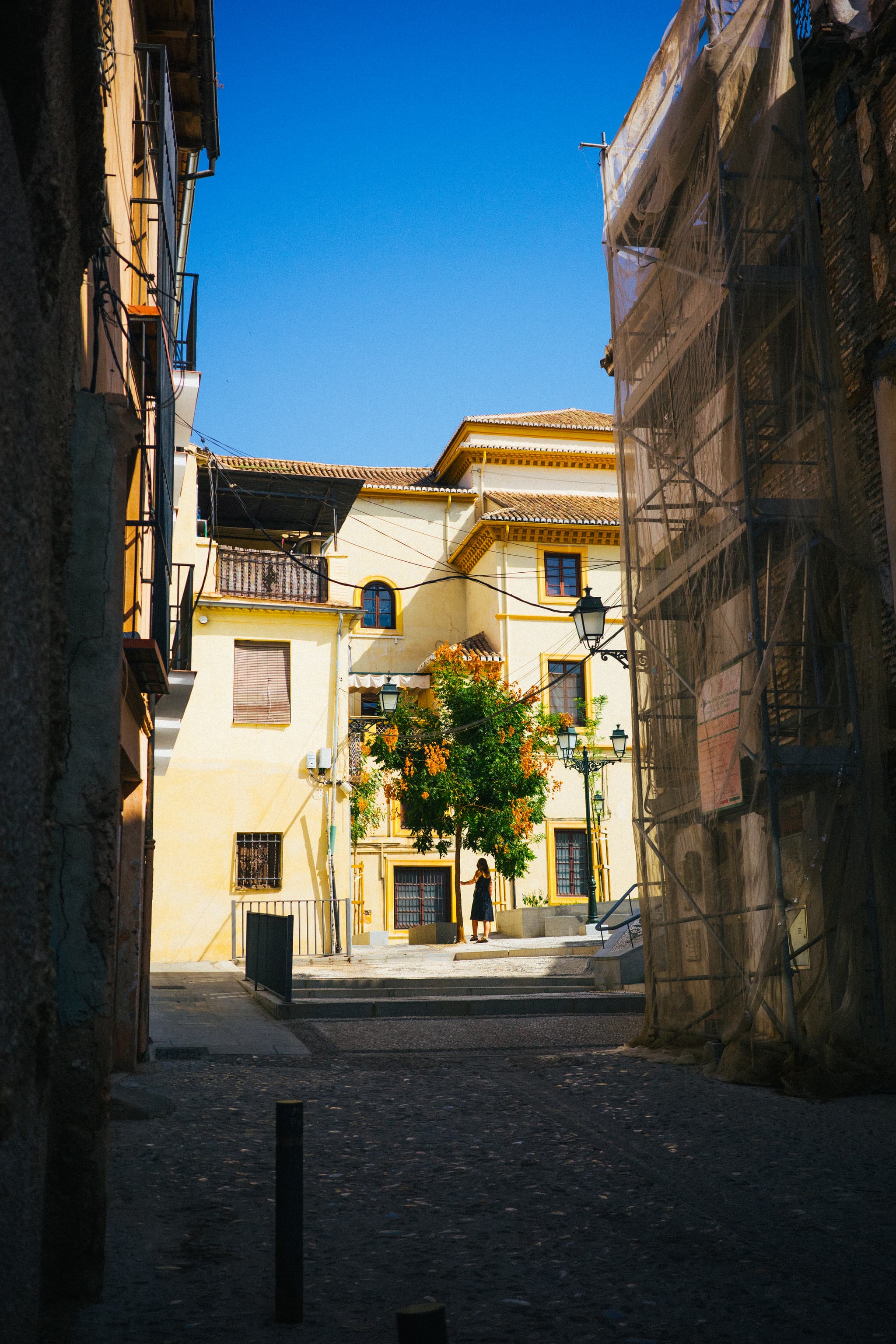 A narrow street leading to a sunlit building with balconies and plants, flanked by other structures under a clear blue sky