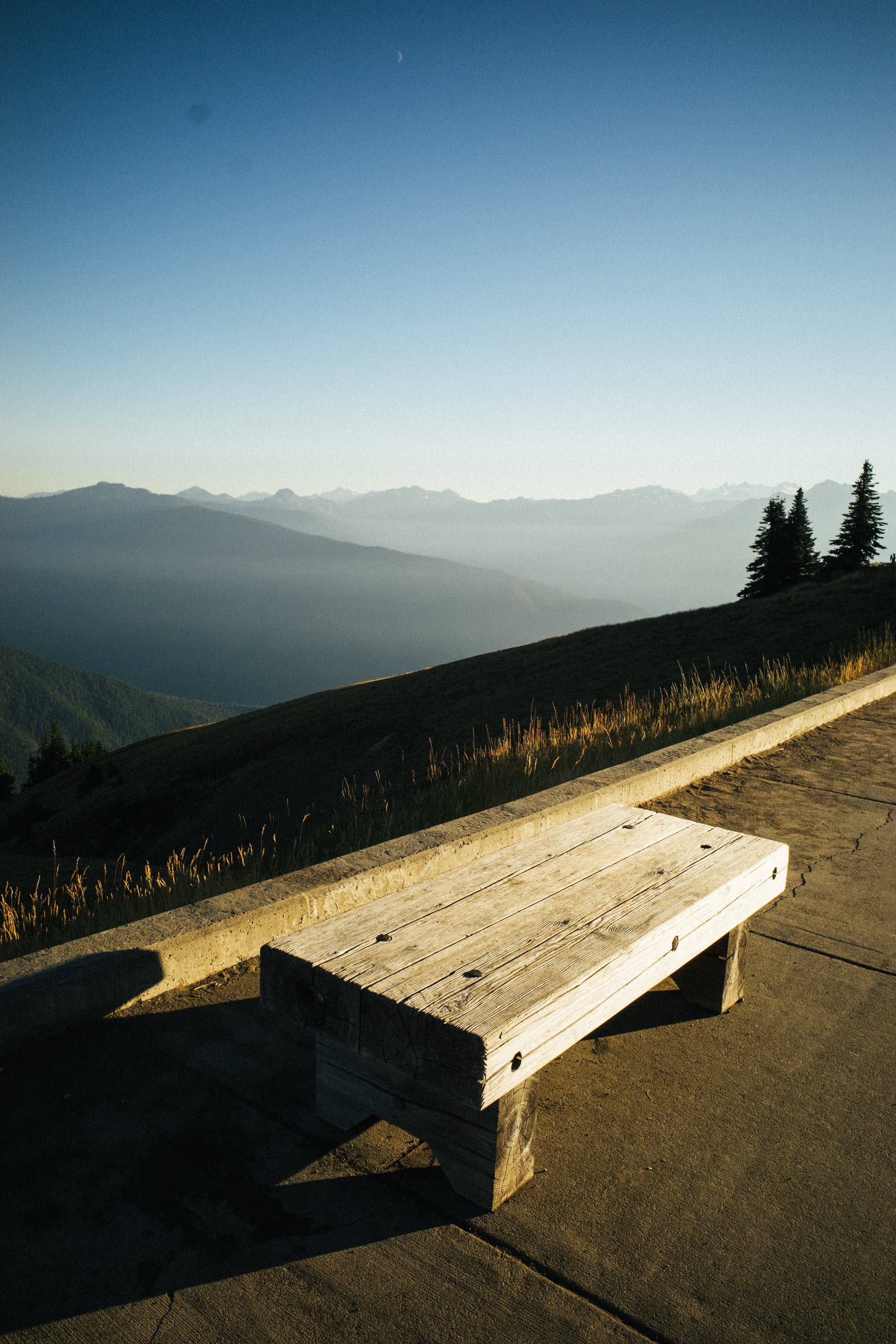 A wooden bench sits at the edge of a paved area with a sweeping view of distant mountains under a clear sky