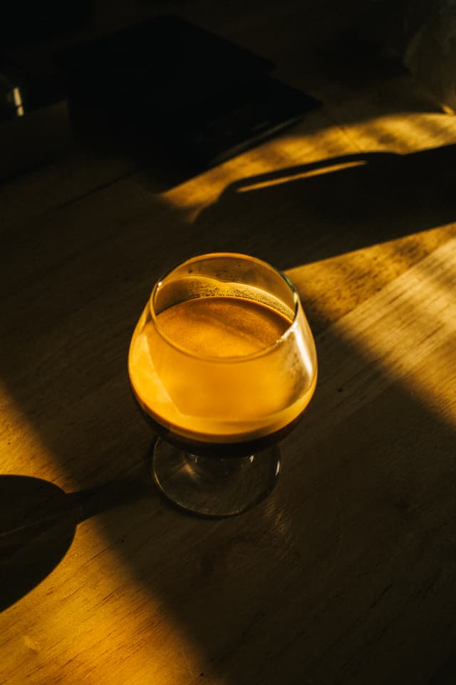 A glass of amber liquid sits on a wooden surface bathed in warm, dramatic lighting, casting a long shadow