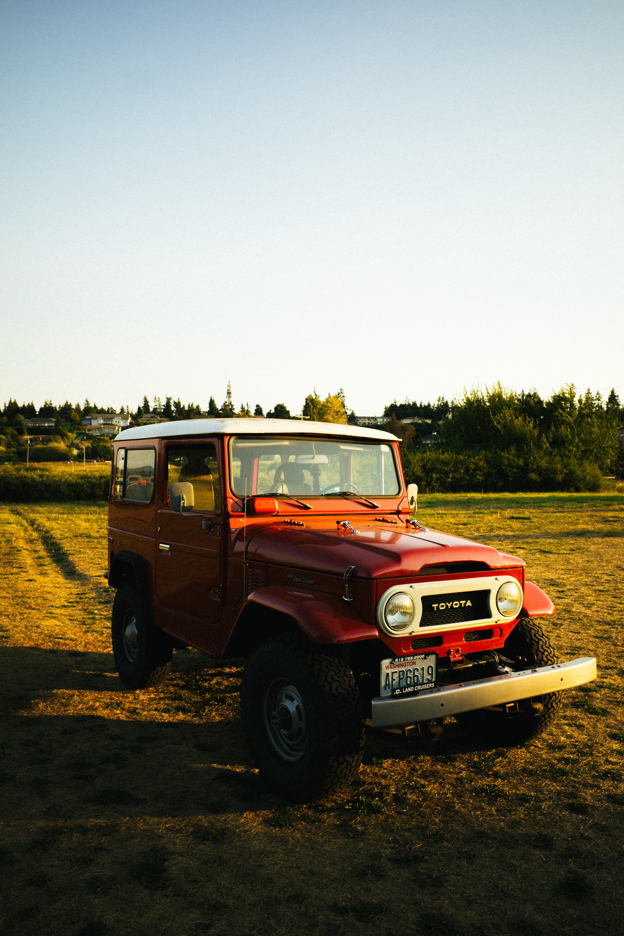 A red vintage off-road vehicle is parked on a grassy field with trees in the background, bathed in the warm glow of sunlight, likely during the late afternoon or early evening