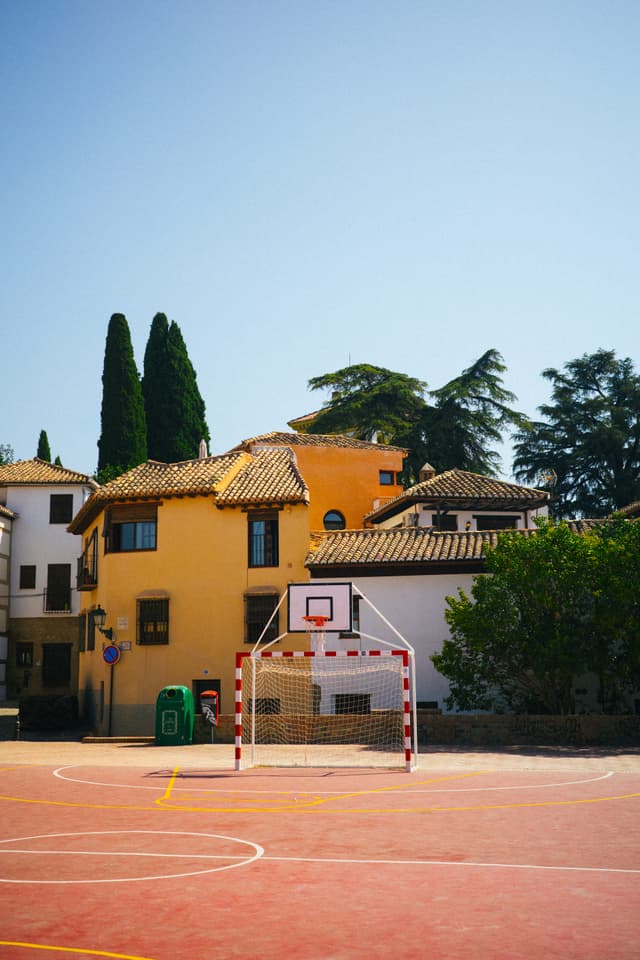 A basketball court with a red surface and white markings in front of a yellow house with a terracotta roof, surrounded by green trees under a clear blue sky