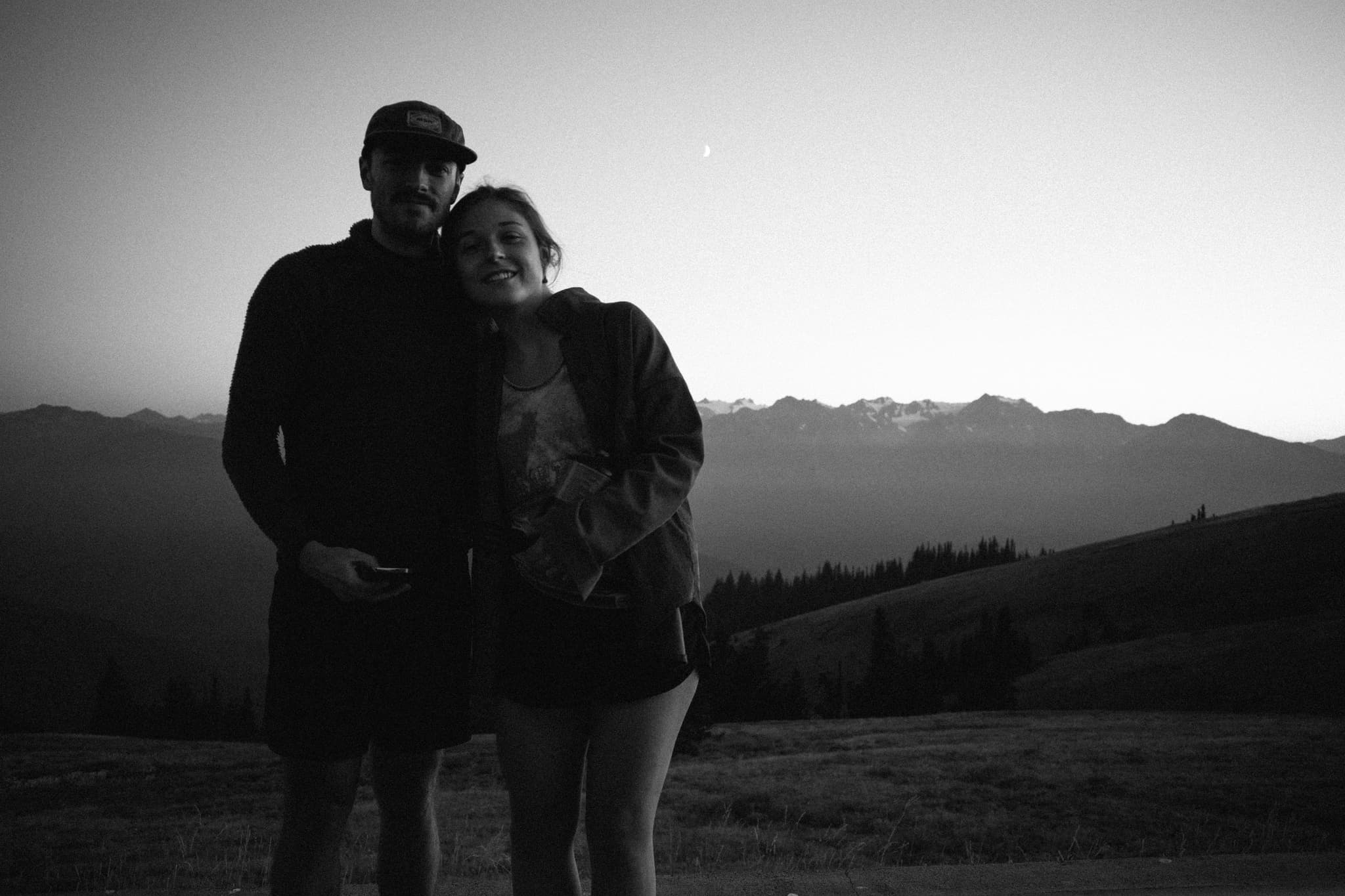 A black and white photo of a couple embracing, with mountains in the background and a twilight sky above