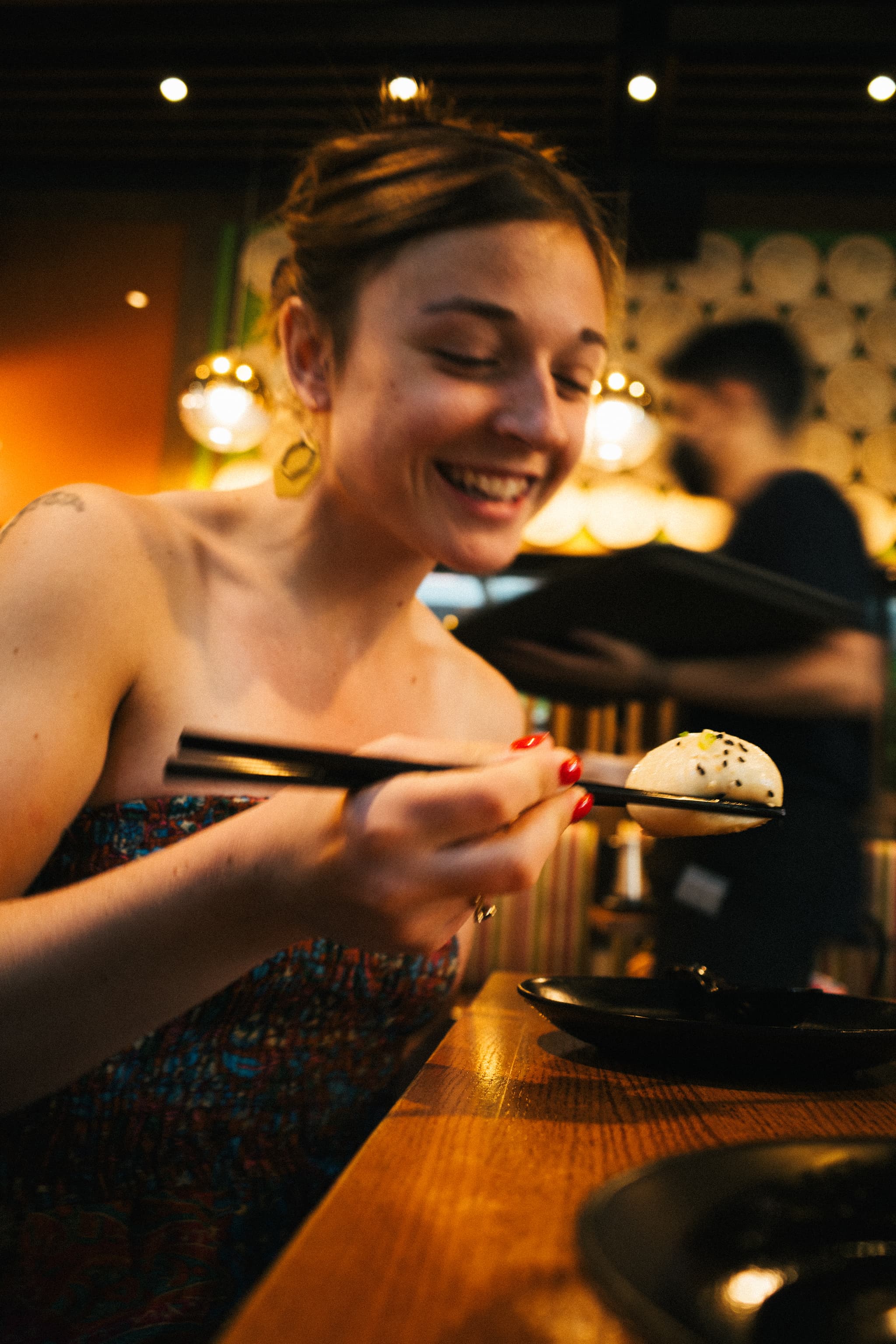 A smiling woman is using chopsticks to hold a piece of sushi, with a blurred restaurant background suggesting a cozy dining atmosphere