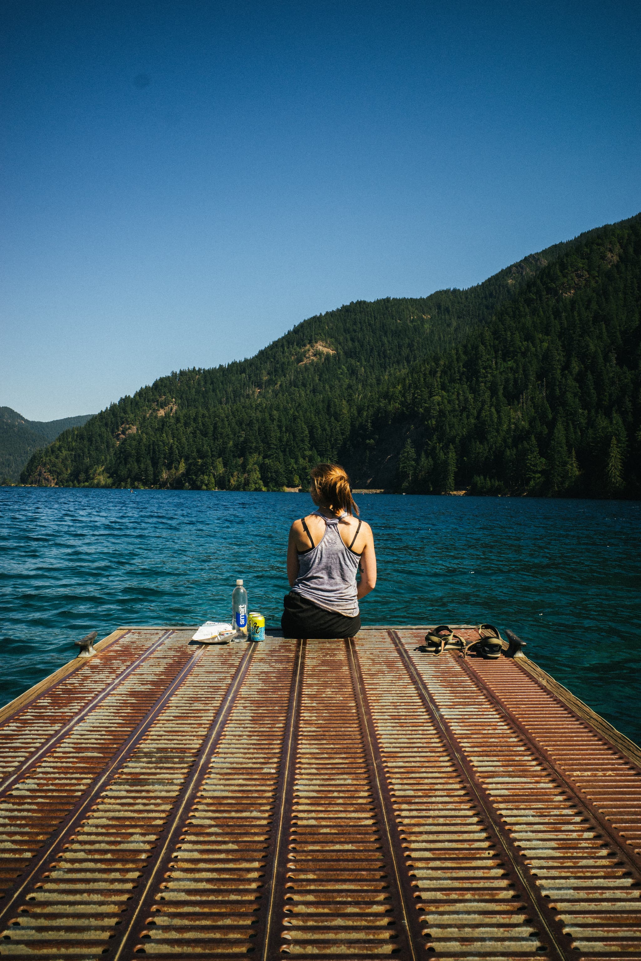 A person sits at the end of a wooden dock overlooking a tranquil blue lake with forested mountains in the background The individual appears to be enjoying a moment of solitude in a serene natural setting