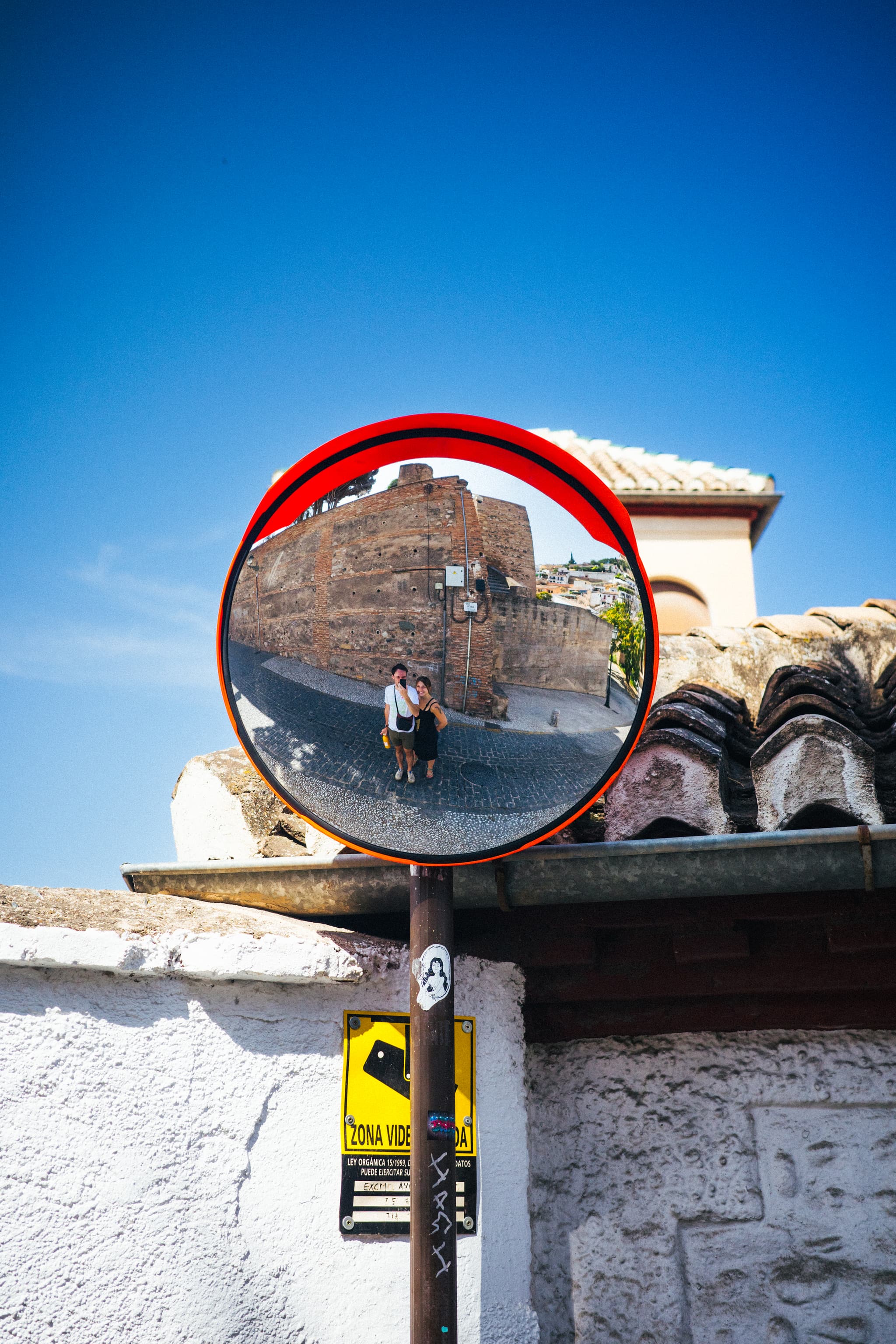 A convex traffic mirror mounted on a pole reflects an image of a person standing in a scenic location with historic buildings under a clear blue sky