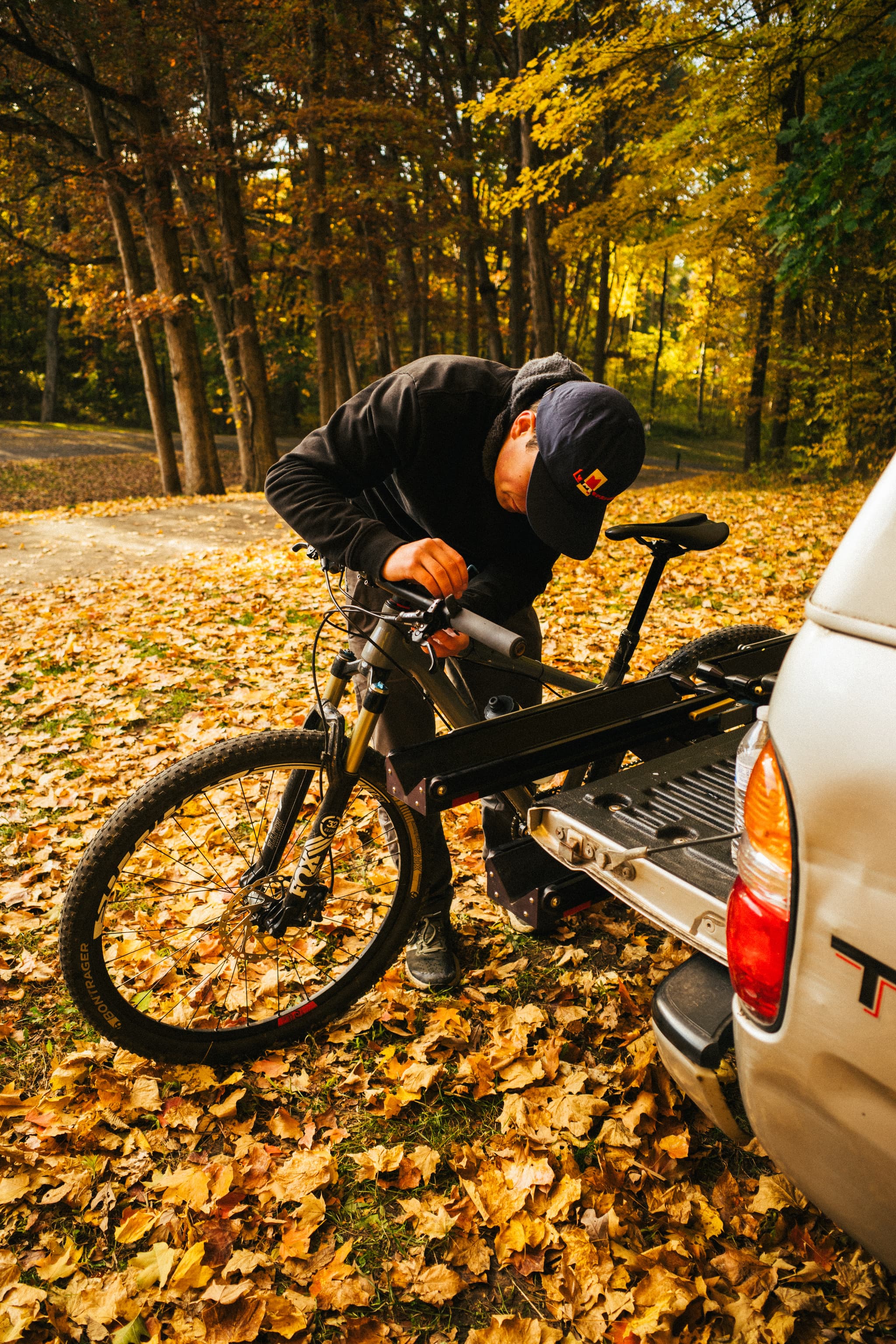 A person is securing a mountain bike onto a rack attached to the back of a car, surrounded by autumn leaves