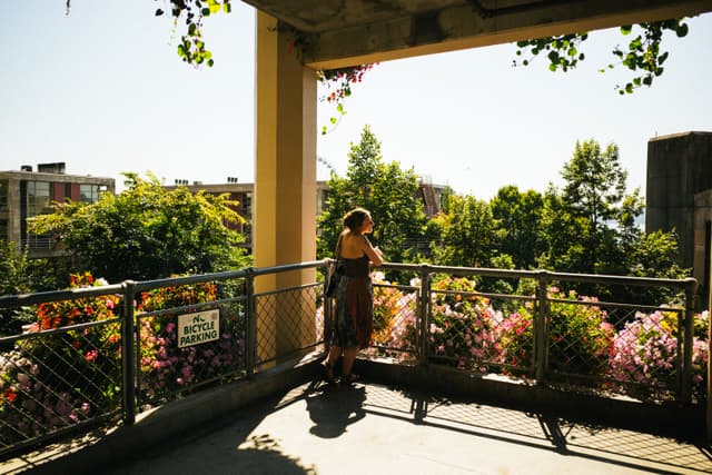 A person stands on a sunlit balcony surrounded by lush greenery and flowers, leaning on the railing and looking out into the distance