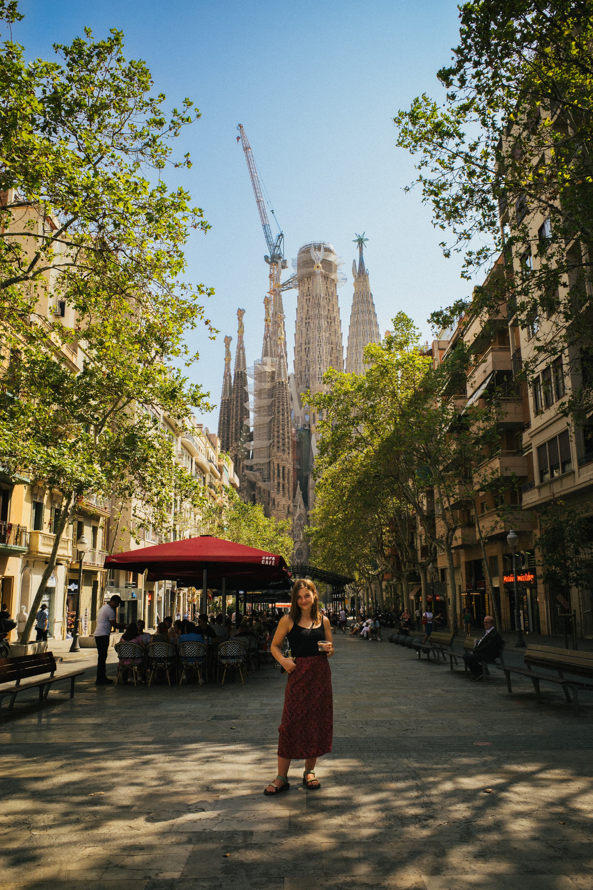 A woman stands on a tree-lined street with the Sagrada Familia cathedral in the background, under a clear blue sky