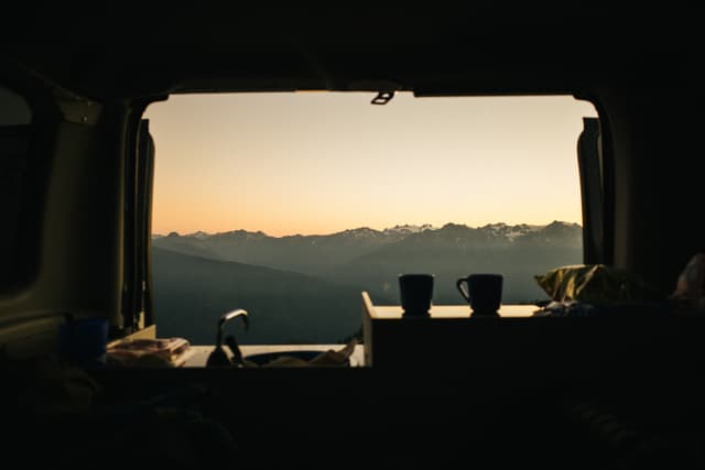 A serene view of a mountainous horizon at dusk or dawn, framed by the open back of a van with silhouetted items and two cups visible inside