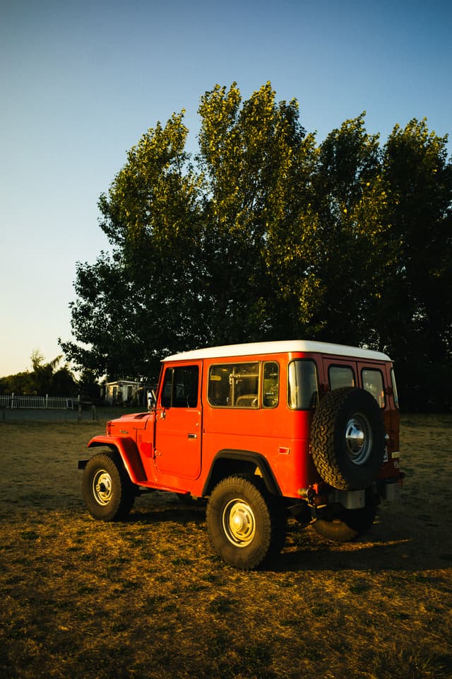 A red off-road vehicle is parked on a grassy field with trees in the background, bathed in the warm light of either sunrise or sunset