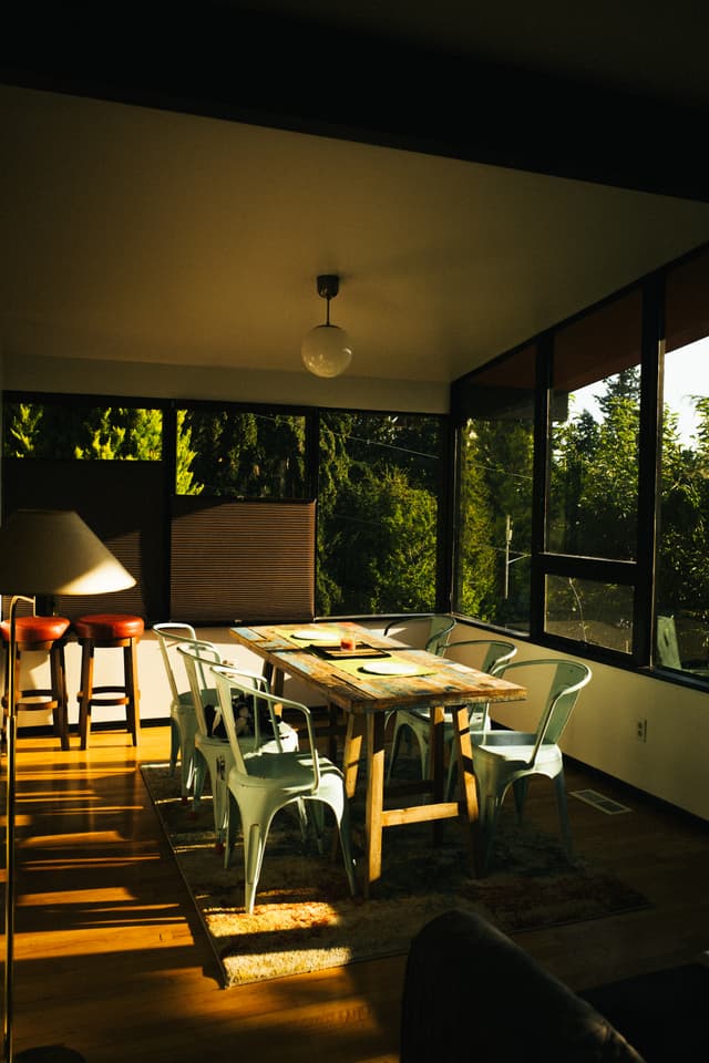 A cozy dining area bathed in warm sunlight with a wooden table, modern chairs, and large windows offering a view of greenery outside