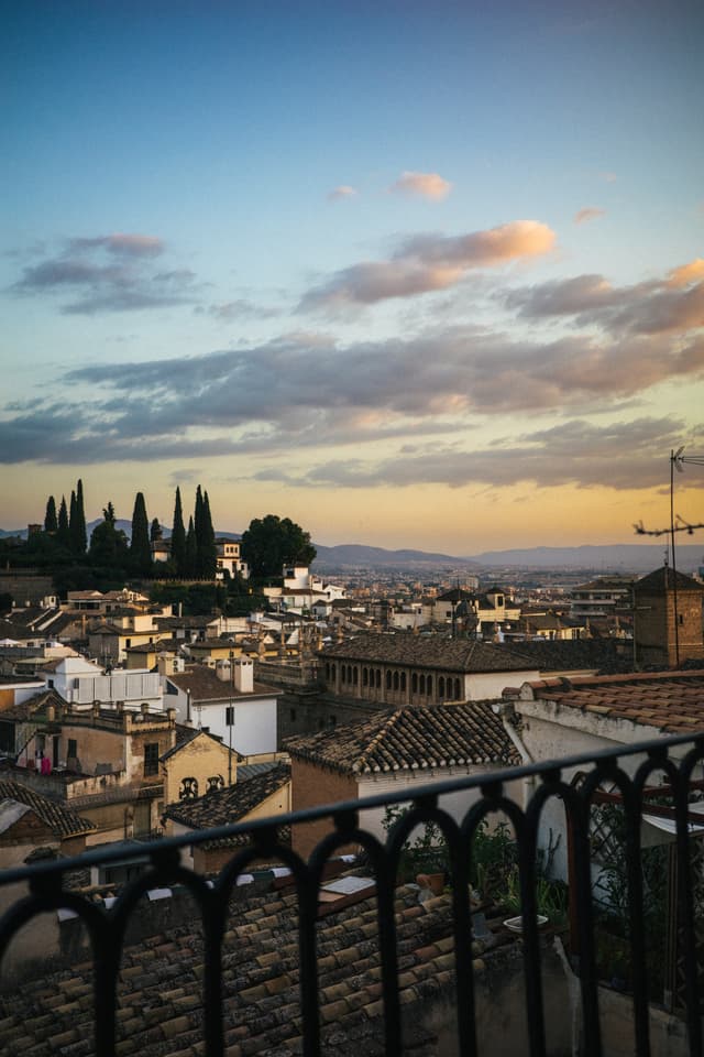 A scenic view of a historic city at dusk, with buildings stretching into the distance, cypress trees on the horizon, and a cloudy sky above, all framed by a wrought iron railing in the foreground