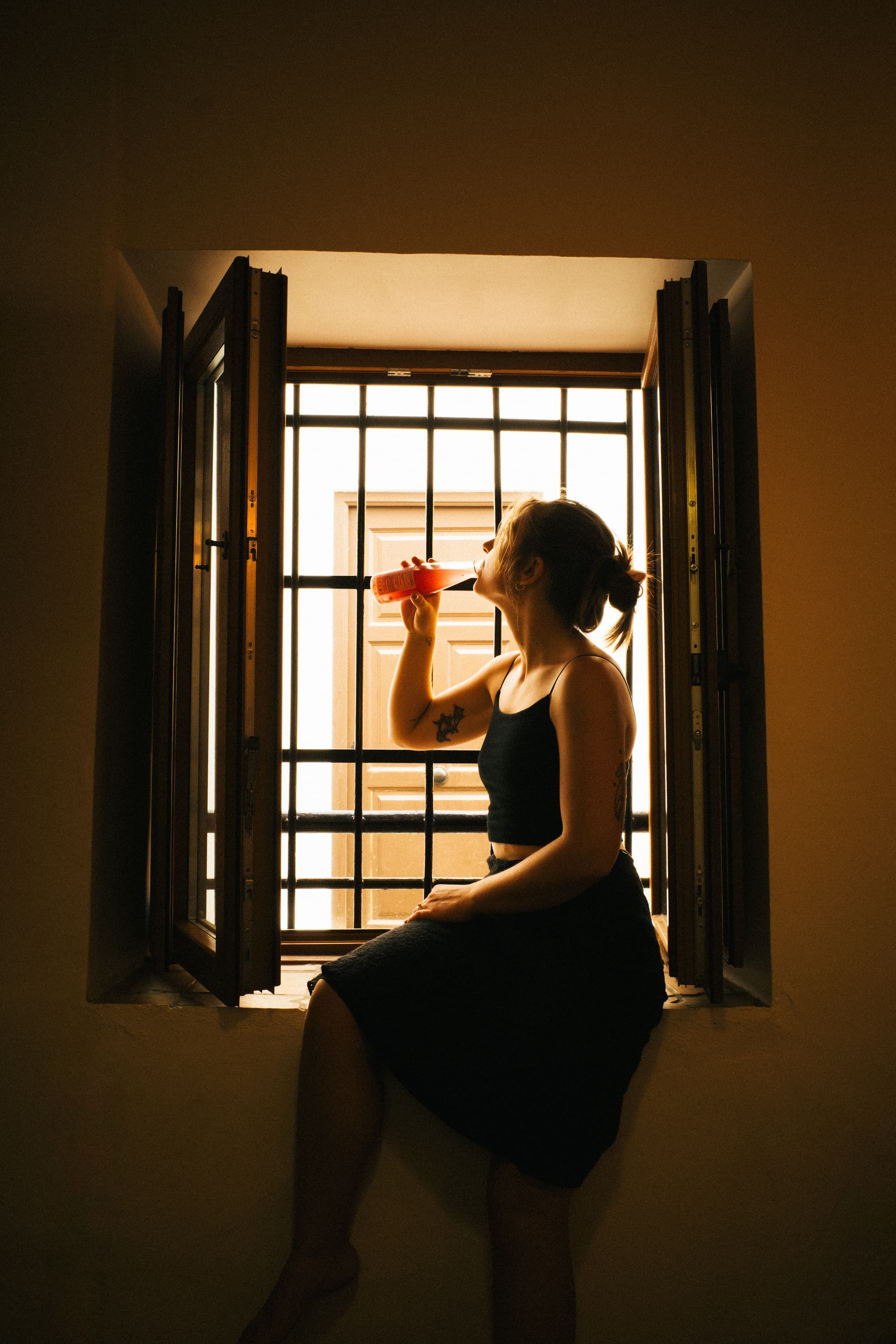 A woman sits by a window with open shutters, sipping from a cup, bathed in soft light