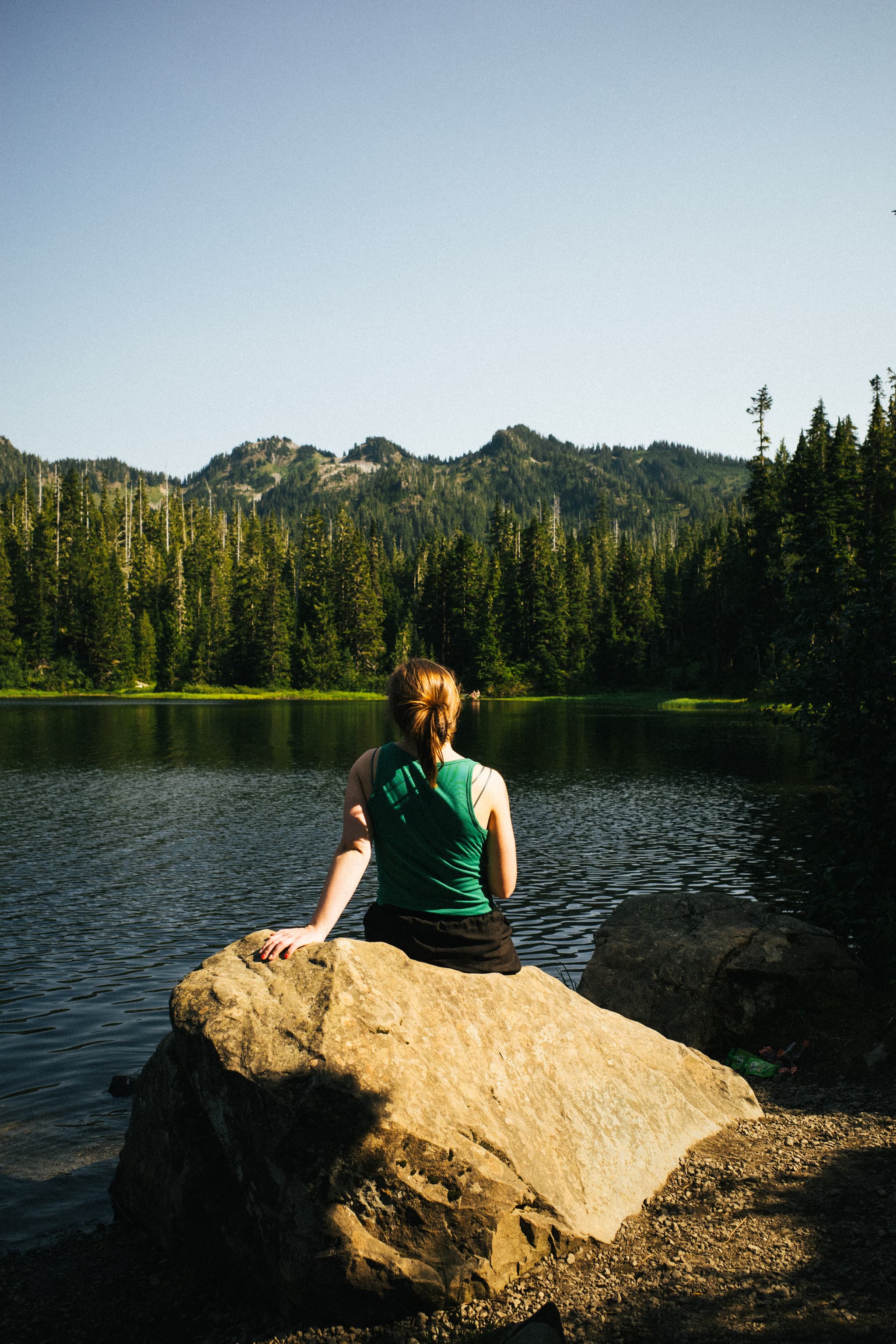 A person sits on a large rock by a tranquil lake, surrounded by forested mountains, gazing at the scenery