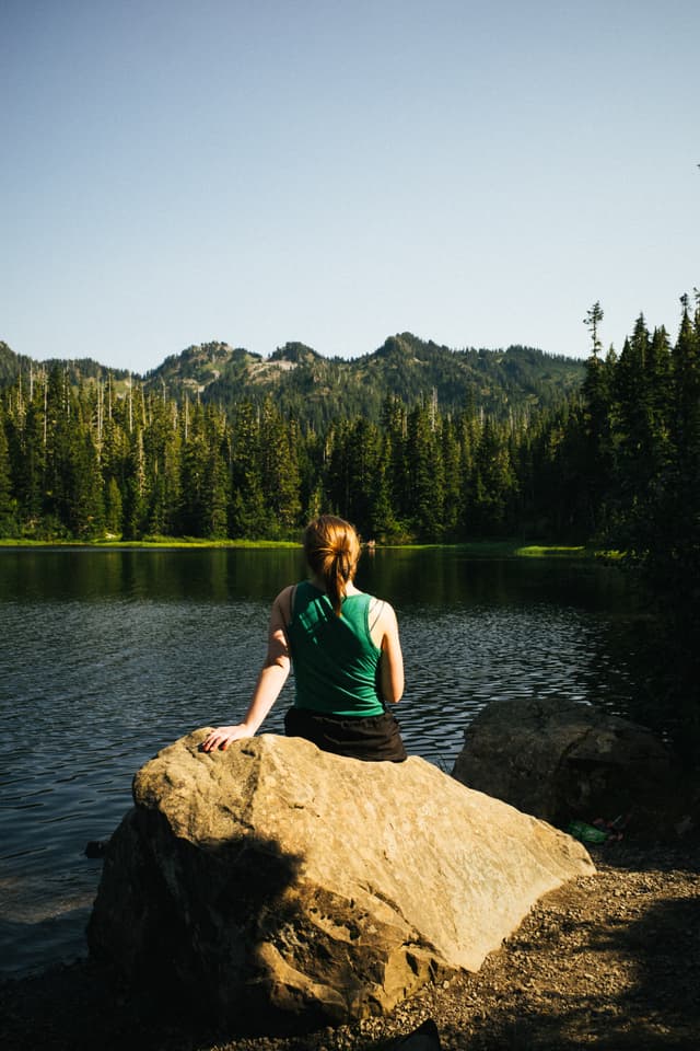 A person sits on a large rock by a tranquil lake, surrounded by forested mountains, gazing at the scenery