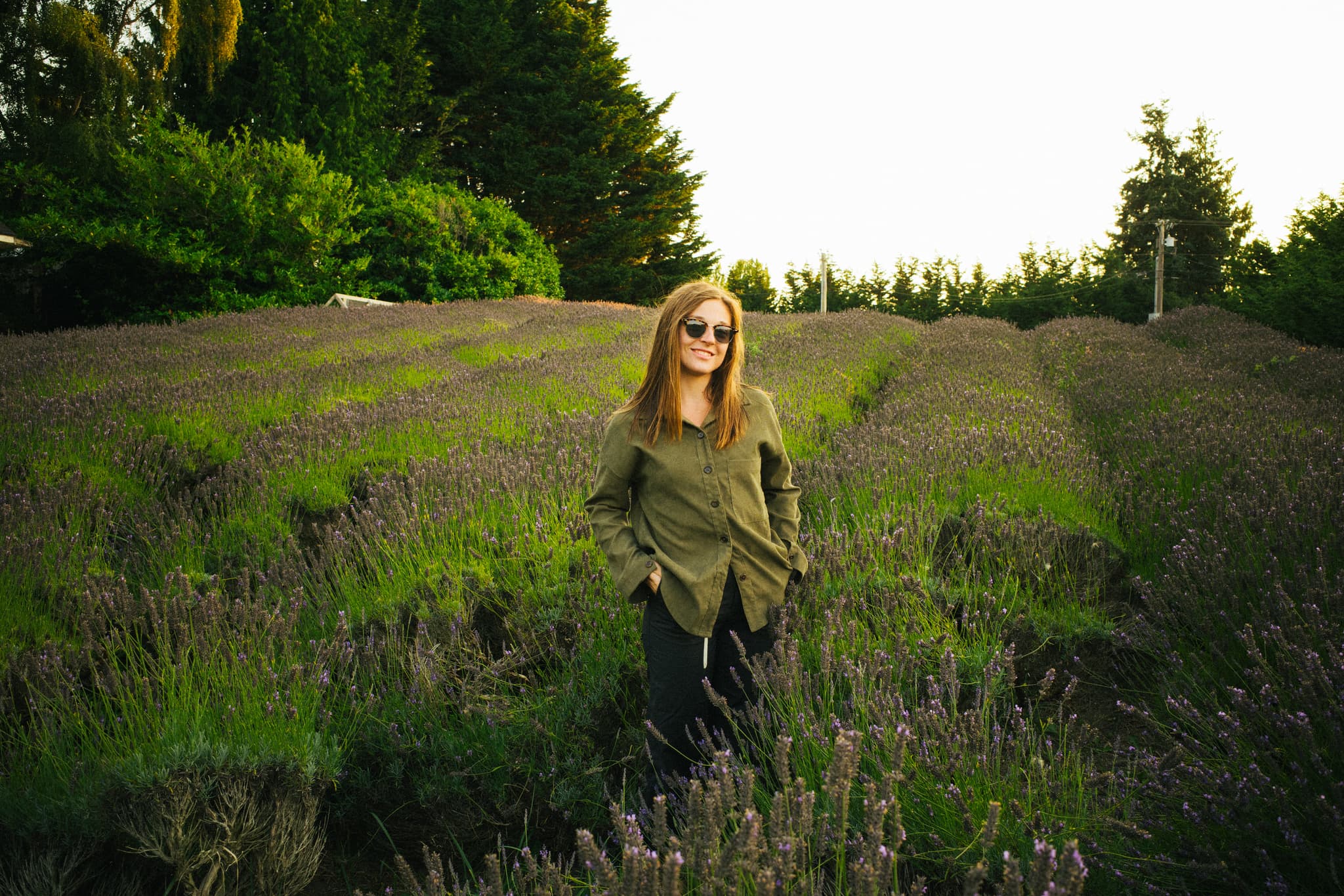 A woman stands in a lavender field, wearing sunglasses and a green jacket, with trees in the background