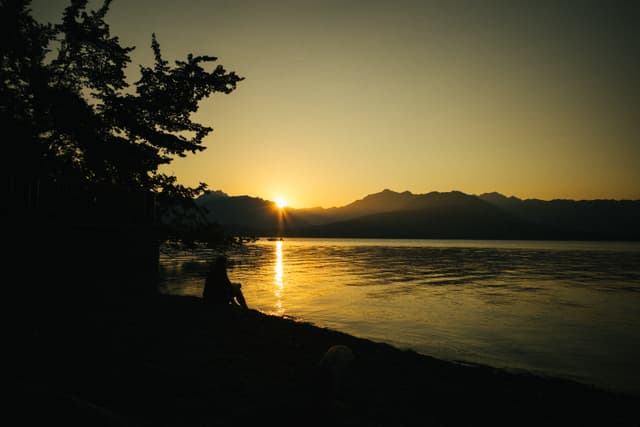 A serene sunset over a body of water with silhouettes of trees and a person on the shore, against a backdrop of distant mountains